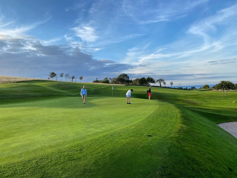 A small group of golfers on a coastal green with the sea beyond.