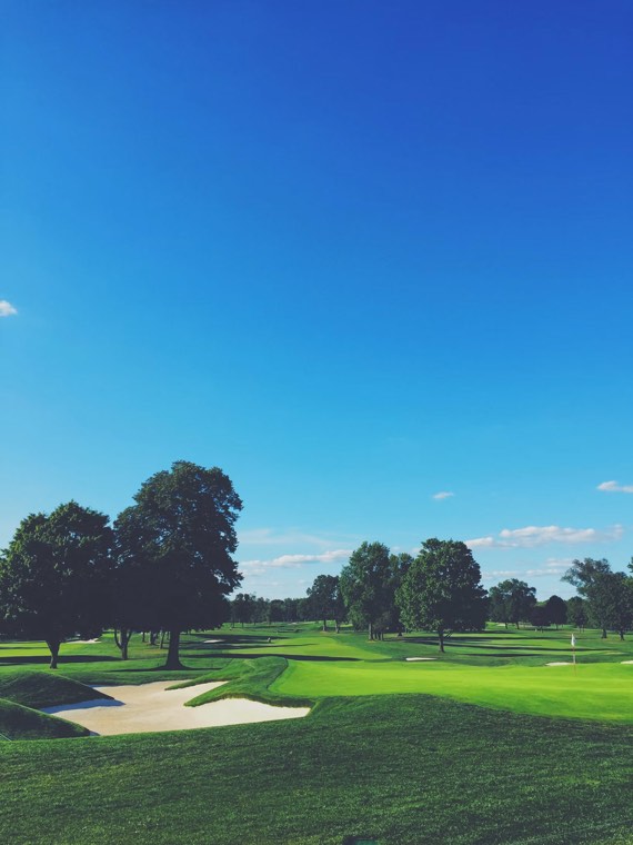 A wide fairway under a clear blue sky, bunkers and mature trees in the distance.