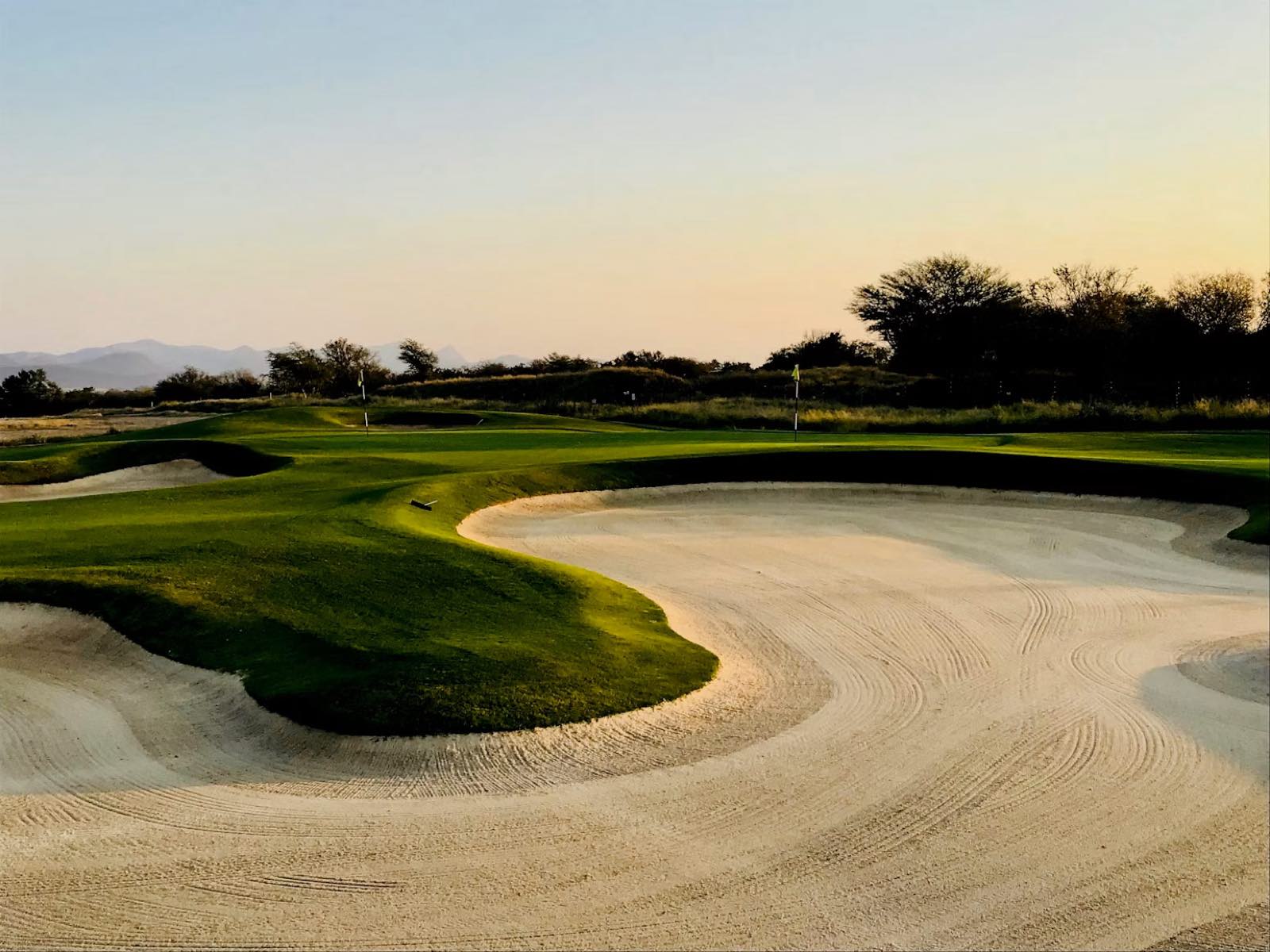 A links-style golf course at dusk, sculpted bunkers curving toward a green.