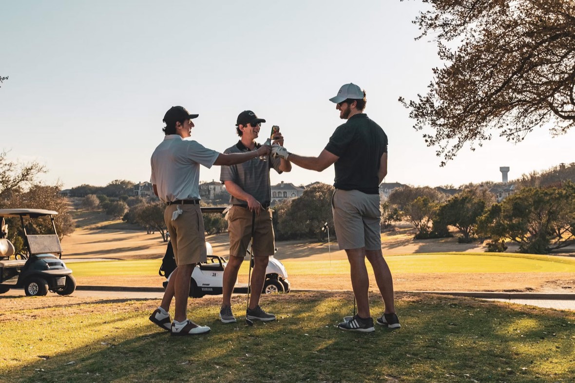 Three golfers standing together on a fairway at golden hour, sharing a drink and laughing.